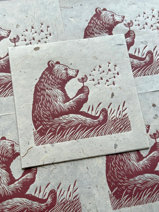 A linocut print featuring a bear sitting in a field of tall grass, gently blowing on a dandelion, with delicate seeds floating in the air. The artwork is rendered in rich reddish-brown tones on textured, light beige paper, showcasing multiple prints of the same design. #Linocut #BearArt #NaturePrint #Dandelion #ArtPrint #WildlifeArt