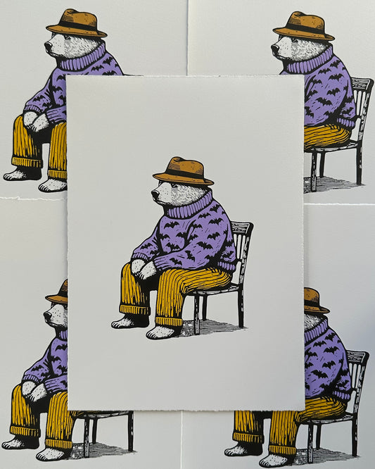A linocut print depicting a seated bear wearing a purple sweater with bat motifs, yellow pants, and a yellow hat. The bear appears contemplative or relaxed, sitting on a black chair against an off-white background.