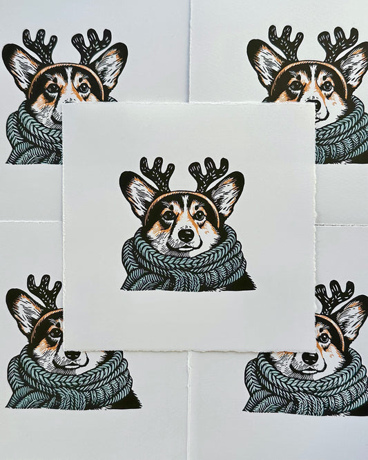 Multi-colour linocut print of a tricolour Pembroke corgi, predominantly black, white, and tan, adorned with an oversized, powder blue knitted scarf wrapped around its neck. The corgi is also wearing a headband with cute antlers, giving it a festive look. The print has a plain white background, which highlights the detailed and colourful depiction of the corgi. Perfect gift for a corgi dog lover and art lovers.
