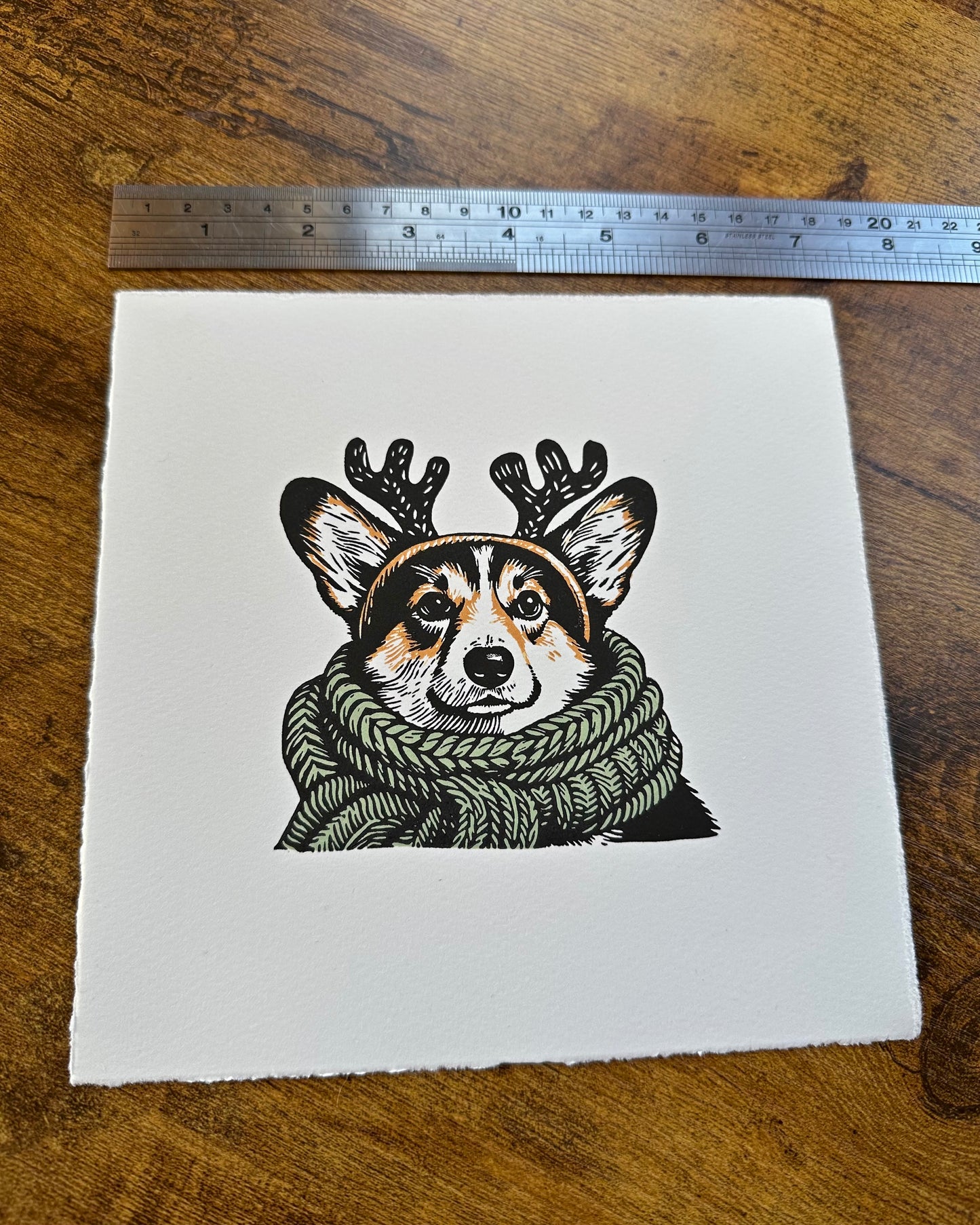 Multi-colour linocut print of a tricolour Pembroke corgi, predominantly black, white, and tan, adorned with an oversized, sage green knitted scarf wrapped around its neck. The corgi is also wearing a headband with cute antlers, giving it a festive look. The print has a plain white background, which highlights the detailed and colourful depiction of the corgi. Perfect gift for a corgi dog lover and art lovers.