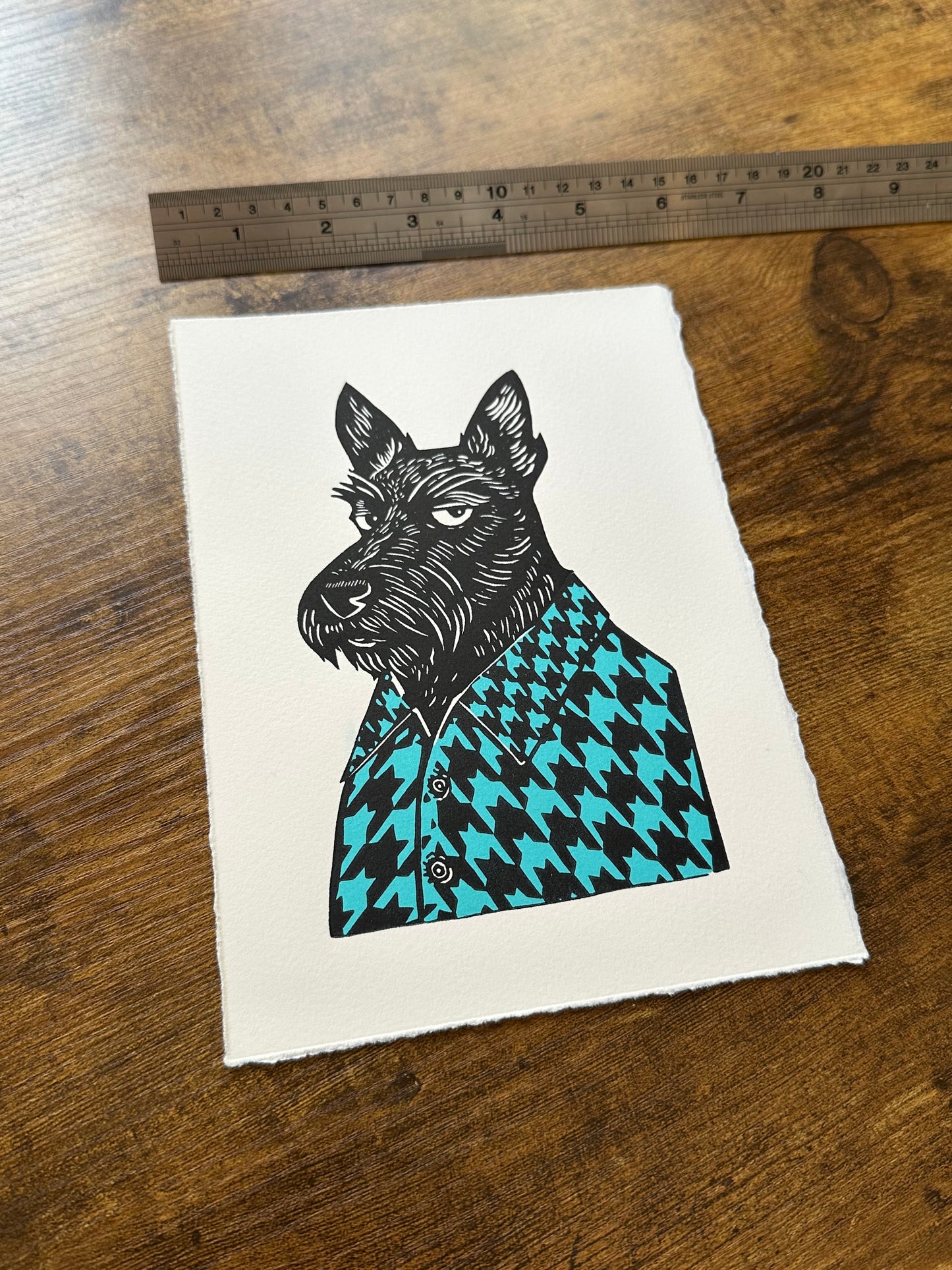 Black and white linocut print of a Scottish terrier wearing a houndstooth-patterned coat in blue, showcasing bold textures and classic design.