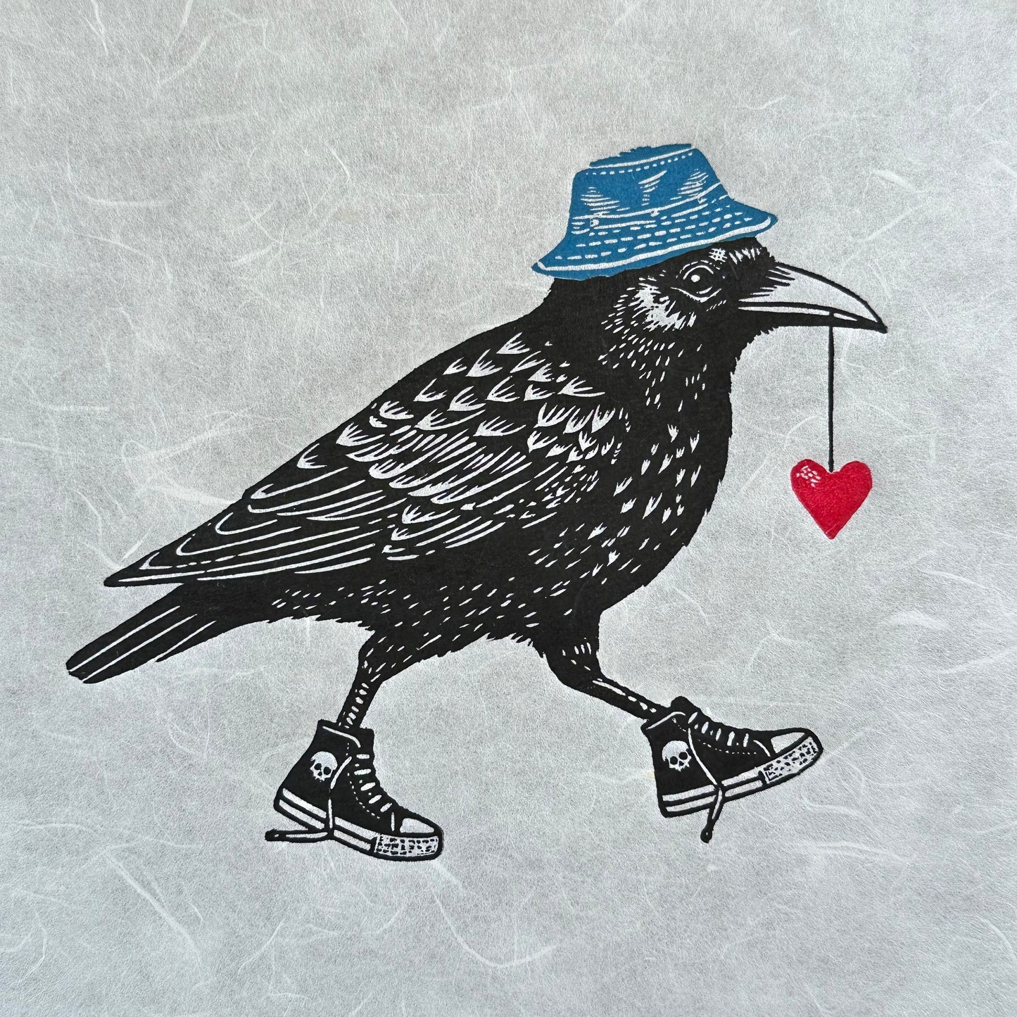 Handmade linocut print of a crow, wearing sneakers with a red heart in its mouth and wearing a blue bucket hat. The crow is detailed with bold, black ink lines, and its kicks/sneakers/trainers add a playful, whimsical touch to the artwork.