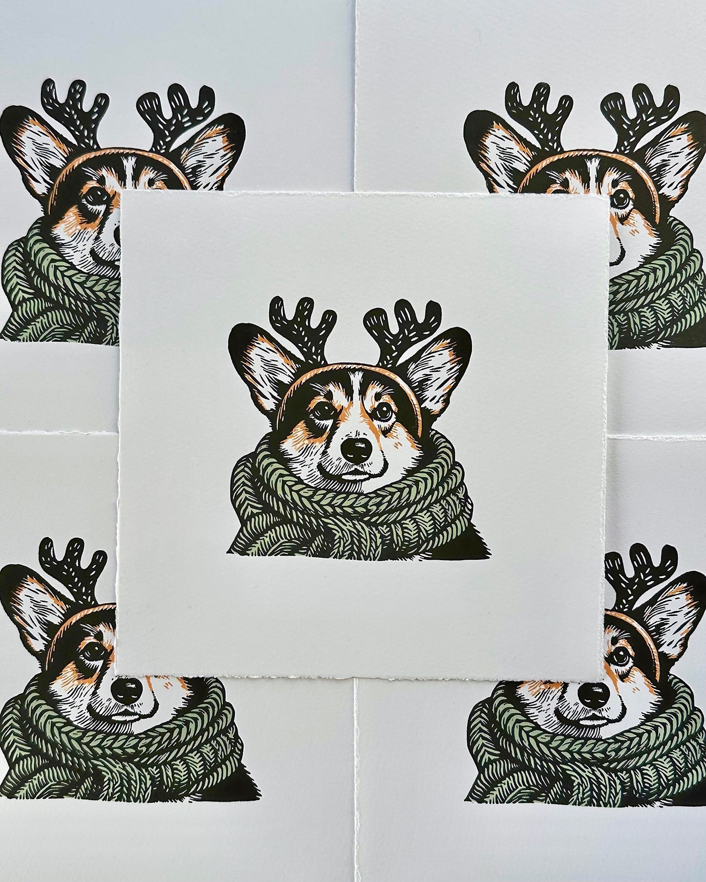 Multi-colour linocut print of a tricolour Pembroke corgi, predominantly black, white, and tan, adorned with an oversized, sage green knitted scarf wrapped around its neck. The corgi is also wearing a headband with cute antlers, giving it a festive look. The print has a plain white background, which highlights the detailed and colourful depiction of the corgi. Perfect gift for a corgi dog lover and art lovers.