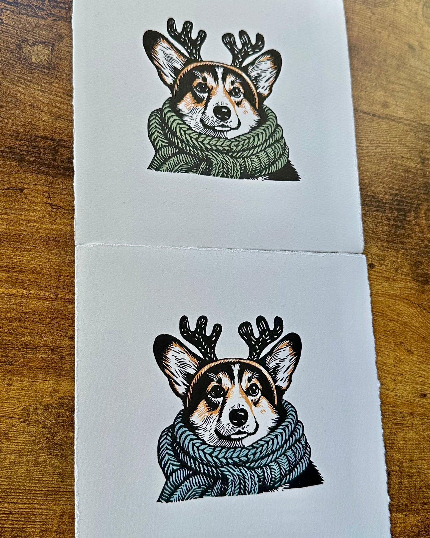 Two multi-colour linocut prints of a tricolour Pembroke corgi, predominantly black, white, and tan, adorned with an oversized, knitted scarf wrapped around its neck. The corgi is also wearing a headband with cute antlers, giving it a festive look. The print has a plain white background, which highlights the detailed and colourful depiction of the corgi. Perfect gift for a corgi dog lover and art lovers.