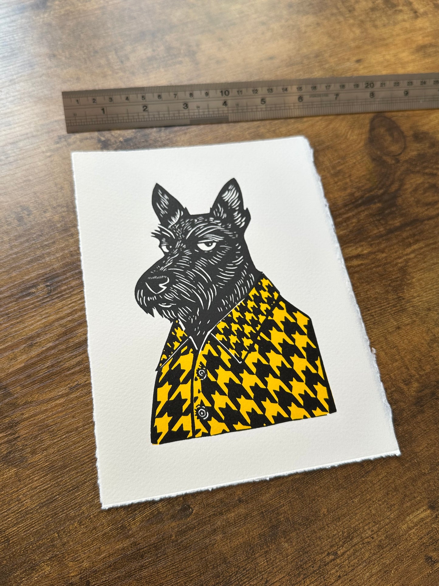Black and white linocut print of a Scottish terrier wearing a houndstooth-patterned coat in yellow, showcasing bold textures and classic design.
