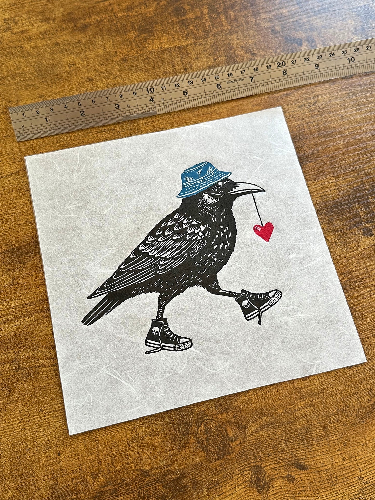 Handmade linocut print of a crow, wearing sneakers with a red heart in its mouth and wearing a blue bucket hat. The crow is detailed with bold, black ink lines, and its kicks/sneakers/trainers add a playful, whimsical touch to the artwork.