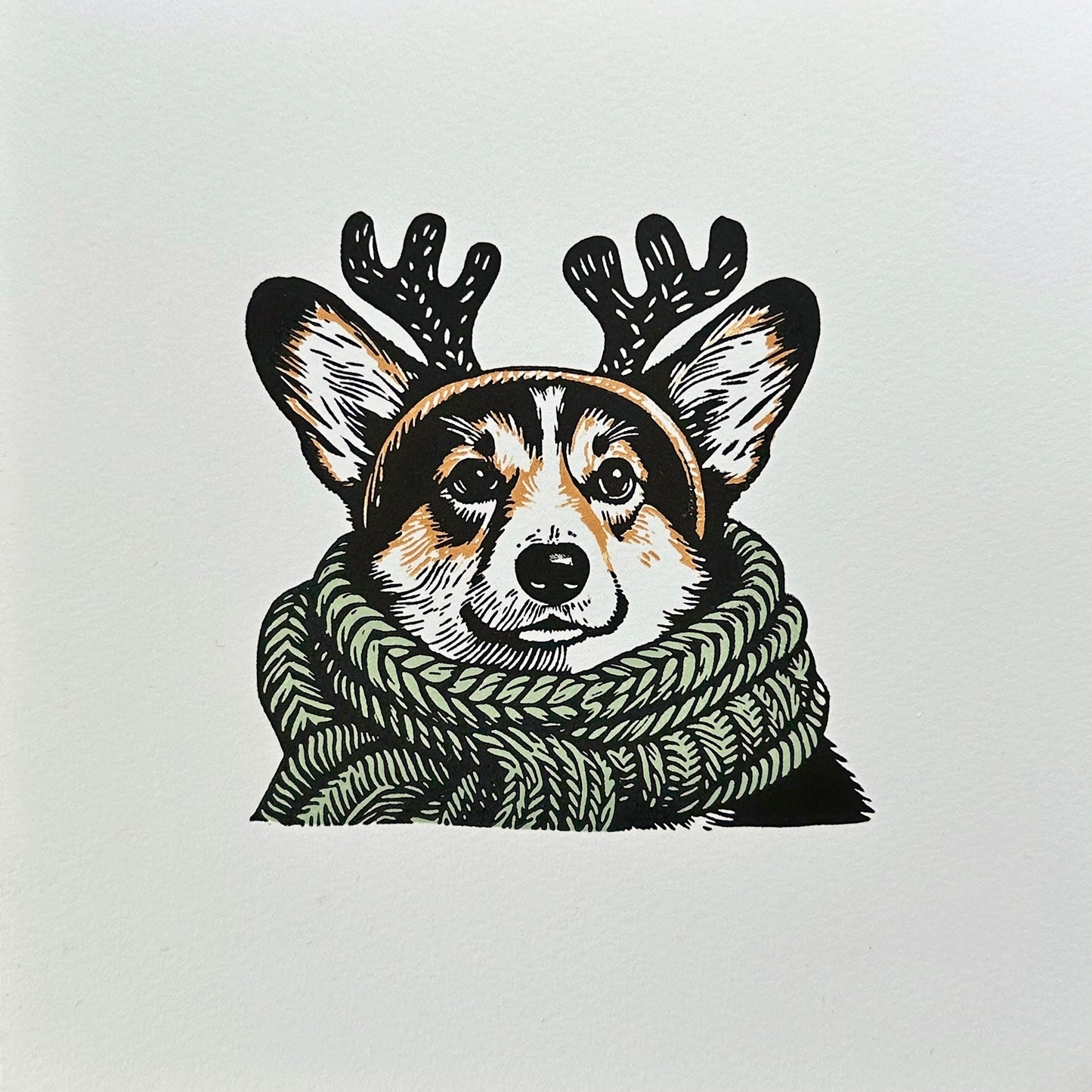 Multi-colour linocut print of a tricolour Pembroke corgi, predominantly black, white, and tan, adorned with an oversized, sage green knitted scarf wrapped around its neck. The corgi is also wearing a headband with cute antlers, giving it a festive look. The print has a plain white background, which highlights the detailed and colourful depiction of the corgi. Perfect gift for a corgi dog lover and art lovers.