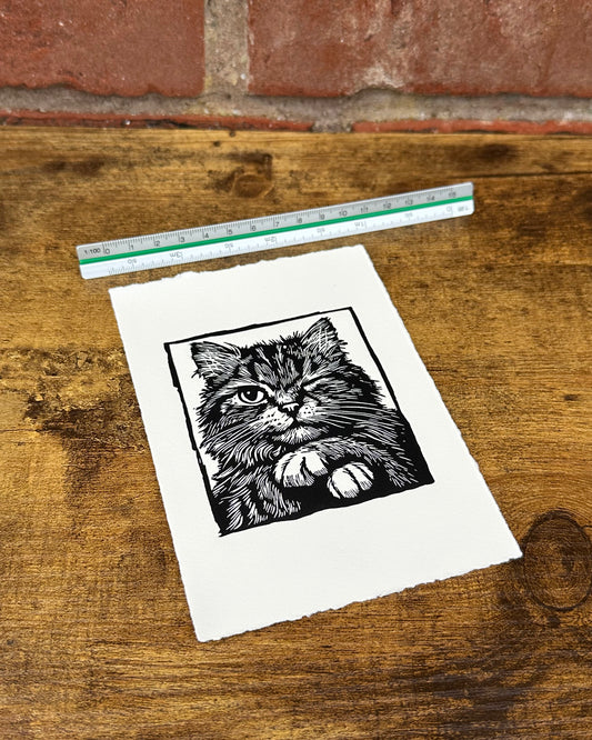 A linocut print of a cat with a ruler for scale on a wooden surface.
