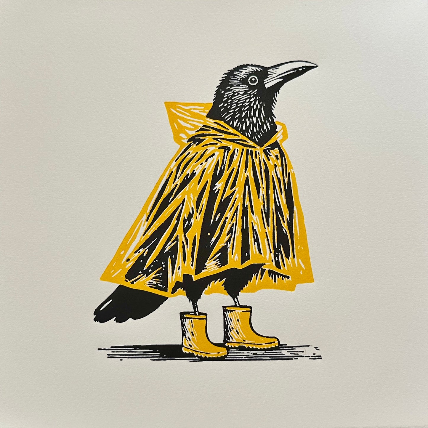 A black crow wearing a yellow raincoat and matching wellington boots. The contrast between the yellow outfit and the bold black crow creates a unique and whimsical image. This handmade linocut print makes a perfect gift for crow and bird lovers.