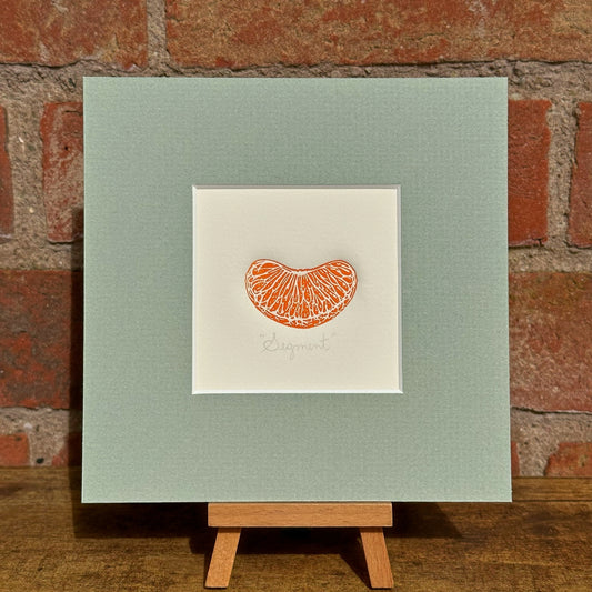 linocut print of an orange segment in a green mount on a wooden easel against a brick wall.