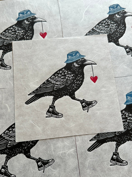 Handmade linocut print of a crow, wearing sneakers with a red heart in its mouth and wearing a blue bucket hat. The crow is detailed with bold, black ink lines, and its kicks/sneakers/trainers add a playful, whimsical touch to the artwork.
