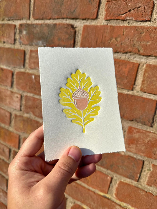 A detailed embossed linocut print of an oak leaf and acorn. The artwork features a delicate layer of ink highlighting the raised textures created by embossing, emphasizing the intricate veins of the oak leaf and the smooth acorn shape. The natural theme celebrates the beauty of oak and acorn with a tactile, handcrafted feel.