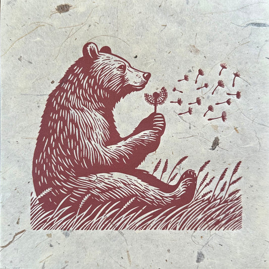 A linocut print featuring a bear sitting in a field of tall grass, gently blowing on a dandelion, with delicate seeds floating in the air. The artwork is rendered in rich reddish-brown tones on textured, light beige paper, showcasing multiple prints of the same design. #Linocut #BearArt #NaturePrint #Dandelion #ArtPrint #WildlifeArt