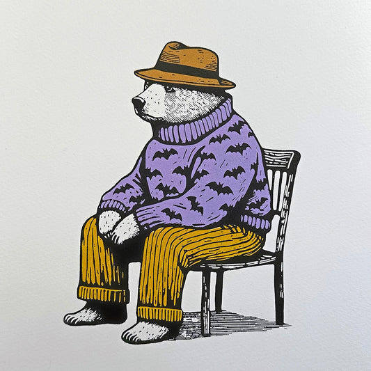 A linocut print depicting a seated bear wearing a purple sweater with bat motifs, yellow pants, and a yellow hat. The bear appears contemplative or relaxed, sitting on a black chair against an off-white background.