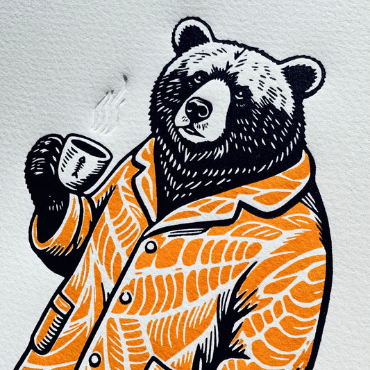 linocut print of a bear wearing orange pyjamas drinking a cup of hot coffee