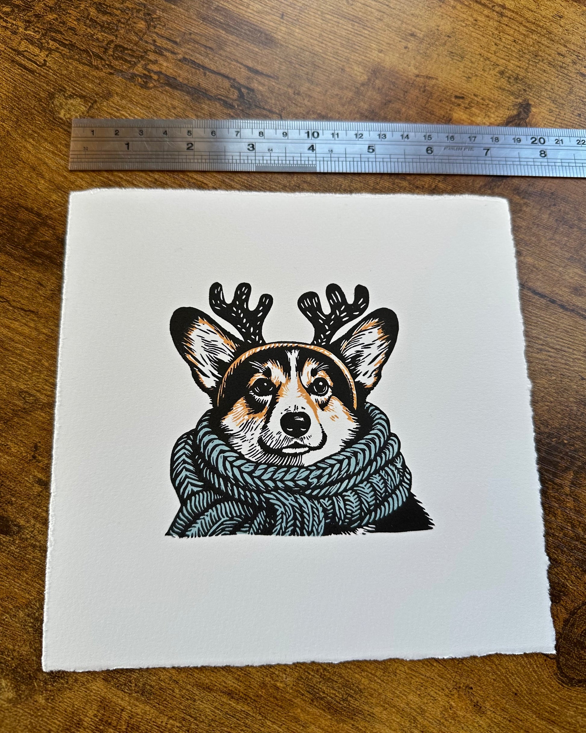 Multi-colour linocut print of a tricolour Pembroke corgi, predominantly black, white, and tan, adorned with an oversized, powder blue knitted scarf wrapped around its neck. The corgi is also wearing a headband with cute antlers, giving it a festive look. The print has a plain white background, which highlights the detailed and colourful depiction of the corgi. Perfect gift for a corgi dog lover and art lovers.