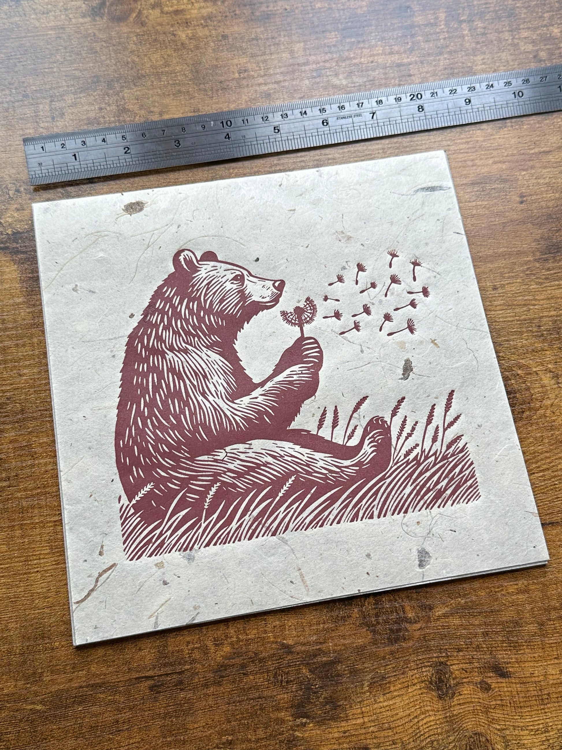 A linocut print featuring a bear sitting in a field of tall grass, gently blowing on a dandelion, with delicate seeds floating in the air. The artwork is rendered in rich reddish-brown tones on textured, light beige paper, showcasing multiple prints of the same design. #Linocut #BearArt #NaturePrint #Dandelion #ArtPrint #WildlifeArt