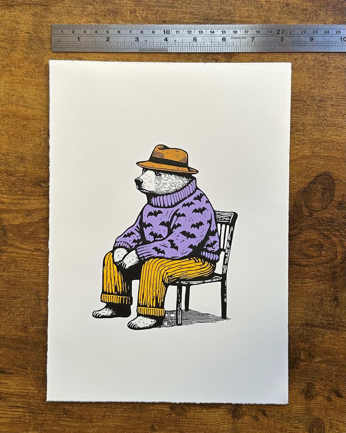 A linocut print depicting a seated bear wearing a purple sweater with bat motifs, yellow pants, and a yellow hat. The bear appears contemplative or relaxed, sitting on a black chair against an off-white background.