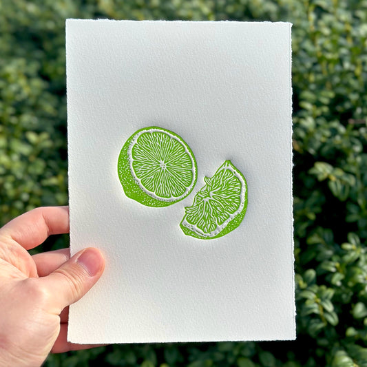 Embossed linocut print of a lime in vibrant green on white paper held against a green leafy background.