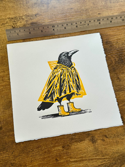 Linocut print of a bird wearing a yellow raincoat and boots on a white background, with a ruler for scale.