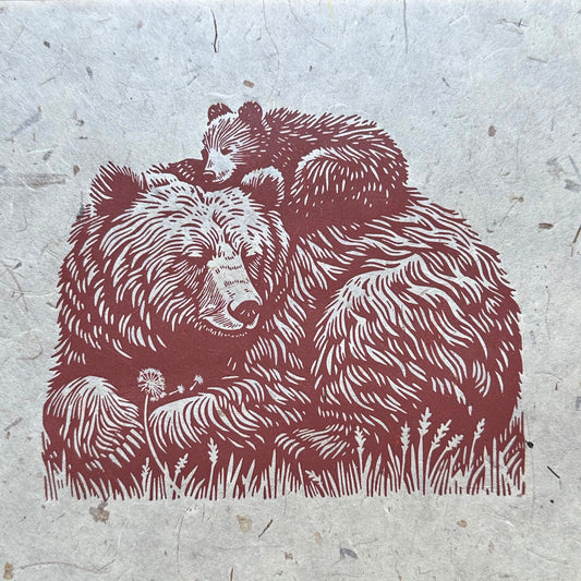Hand-carved linocut print of a mother bear and her cub on textured paper, lying in a grassy field with a dandelion and floating seeds. Nature-inspired animal artwork for home or wall décor.