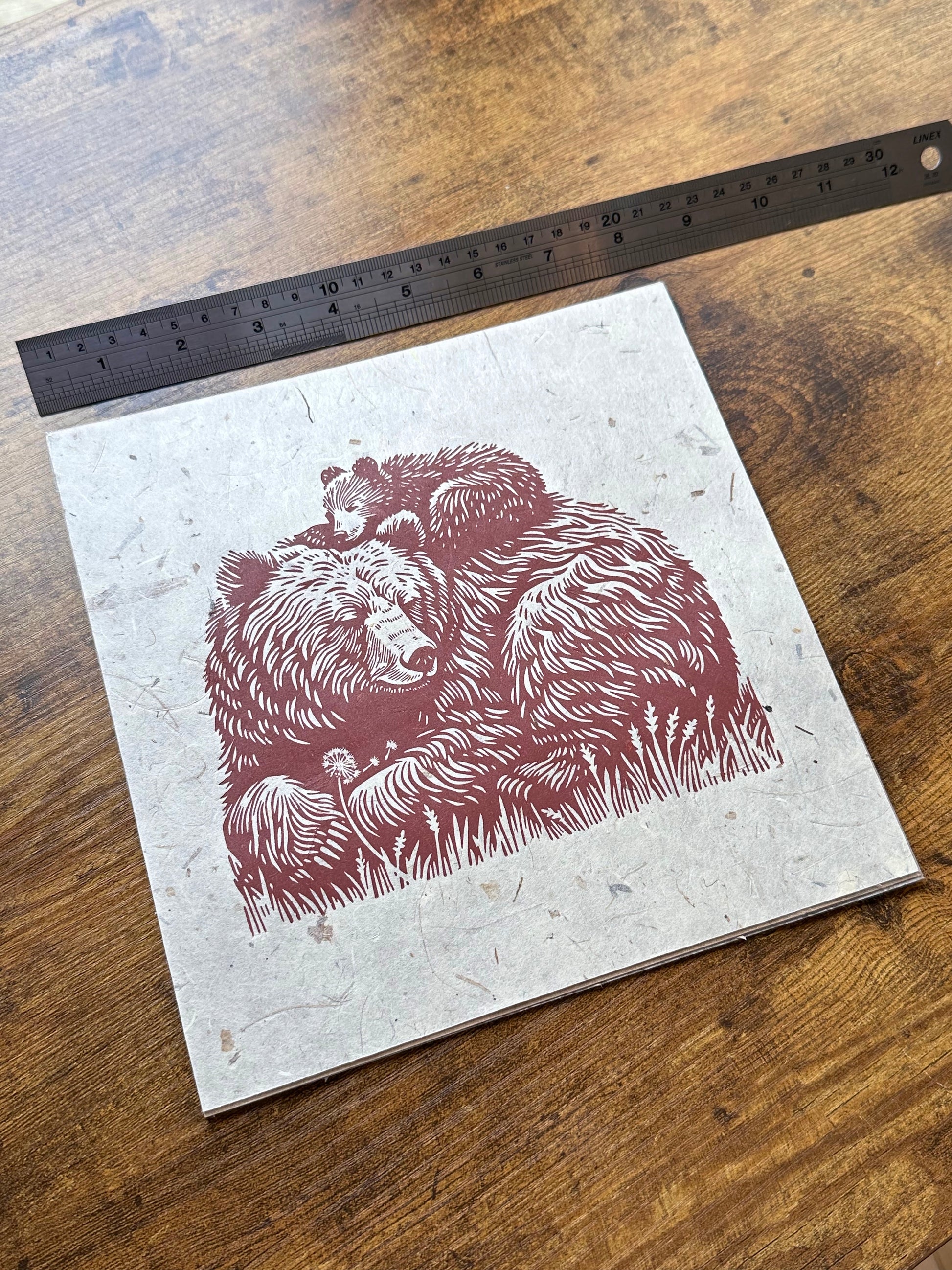 Linocut print of a mother bear and her cub on textured paper with a ruler for scale on a wooden surface. 