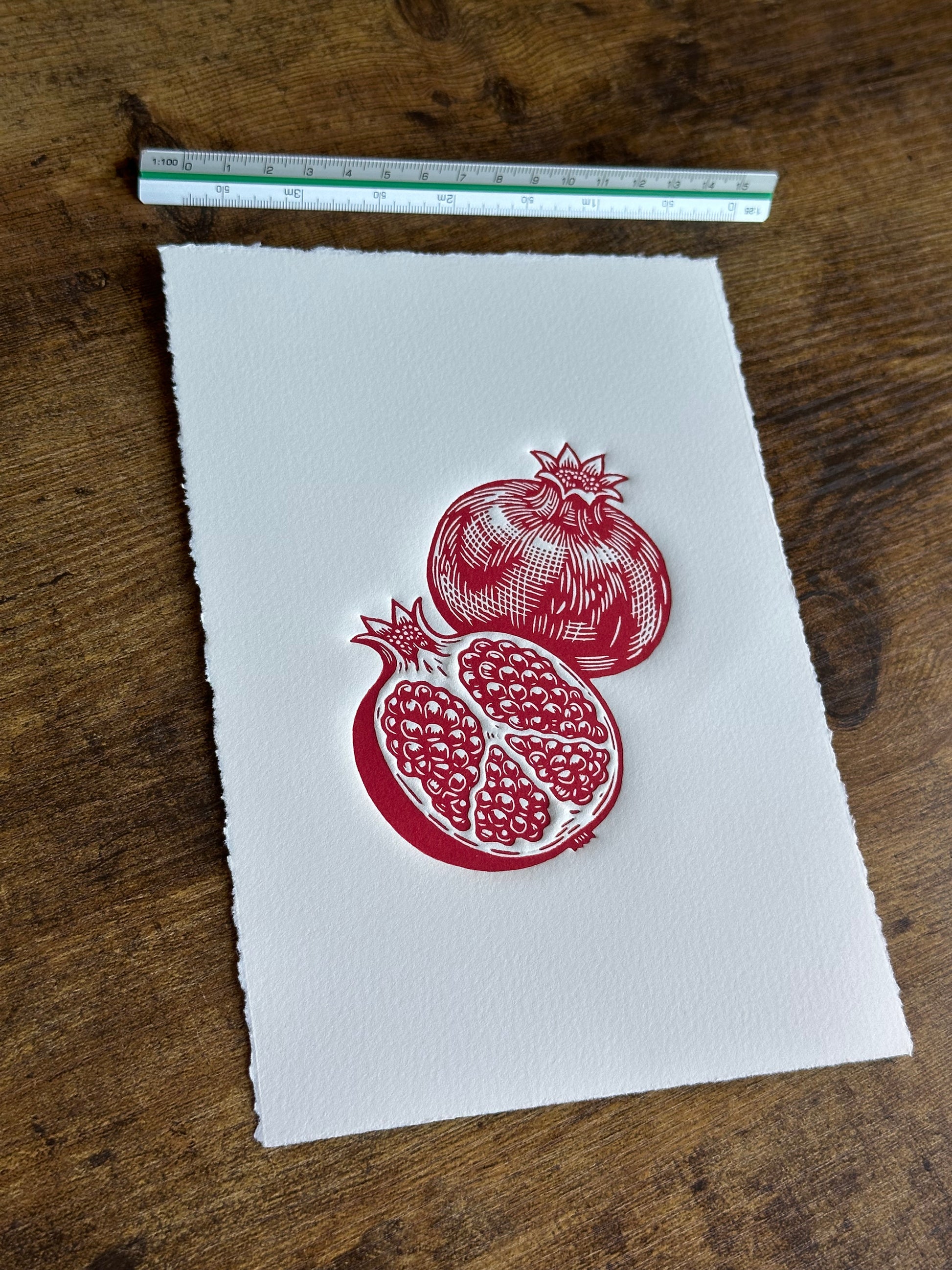 A linocut print with two pomegranates, placed on a brown wooden surface next to a small ruler for scale.