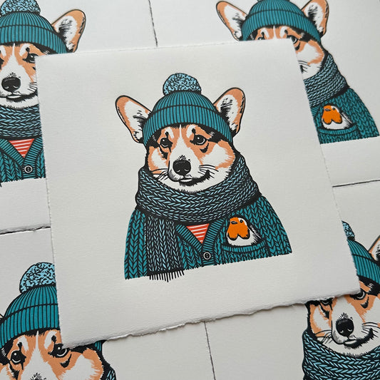 Linocut print of a corgi wearing a bobble hat with a scarf and cardigan. A robin is nestled in the pocket, bright colours