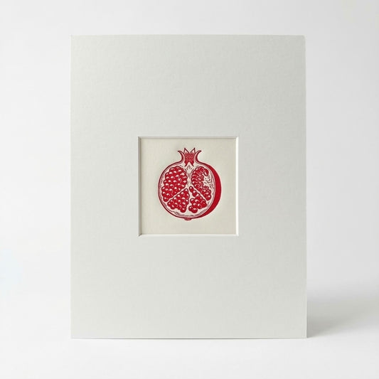 A linocut print featuring a red pomegranate illustration centred within a wide white mounted board, photographed against a clean white background.
