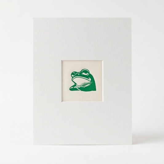 A mounted linocut print with a centred inset showing a green frog illustration on an off white background, photographed against a clean white backdrop.