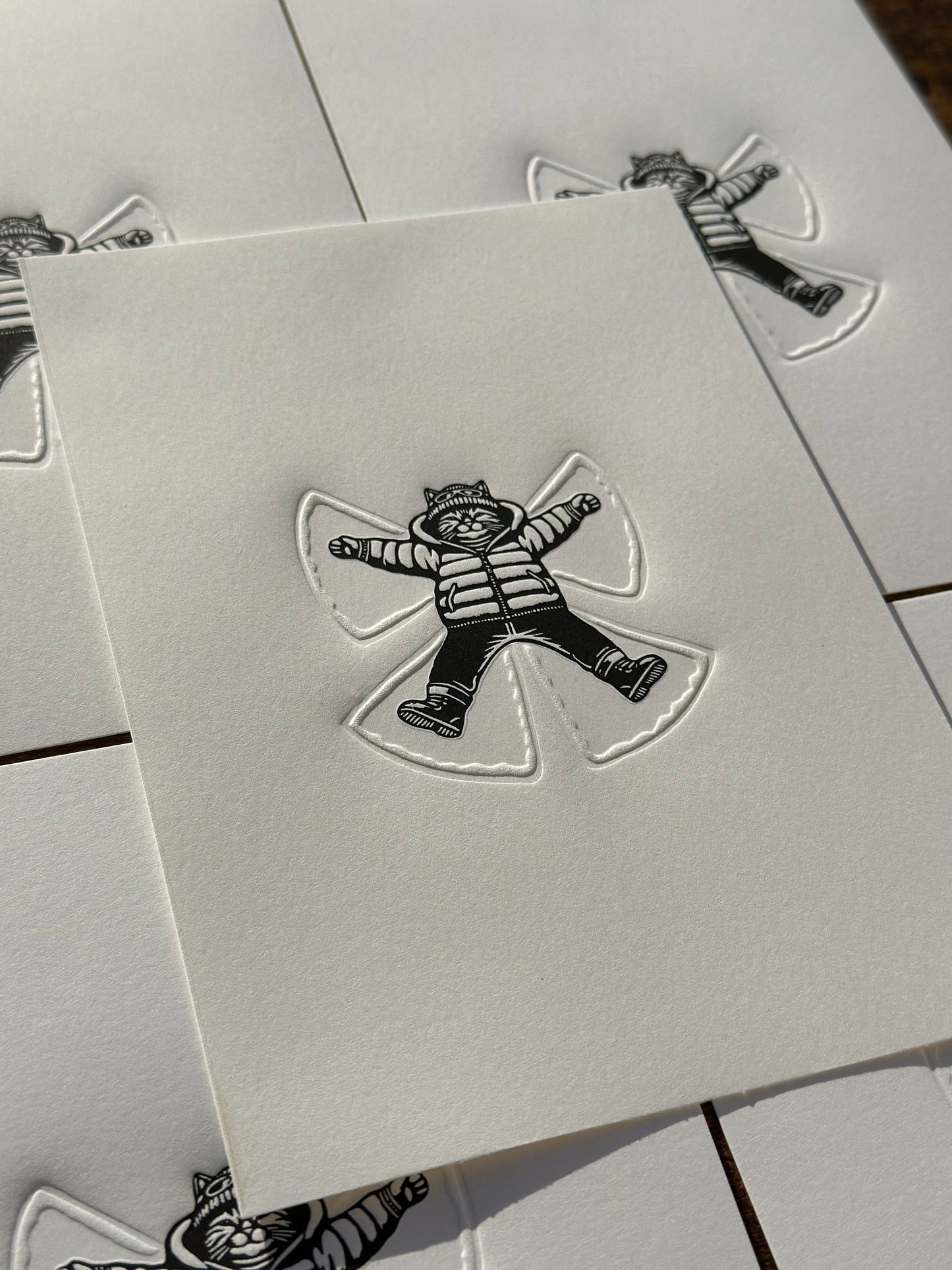 a black and white linocut print of a cat making a snow angel in the snow, the angel is embossed into the paper. 

