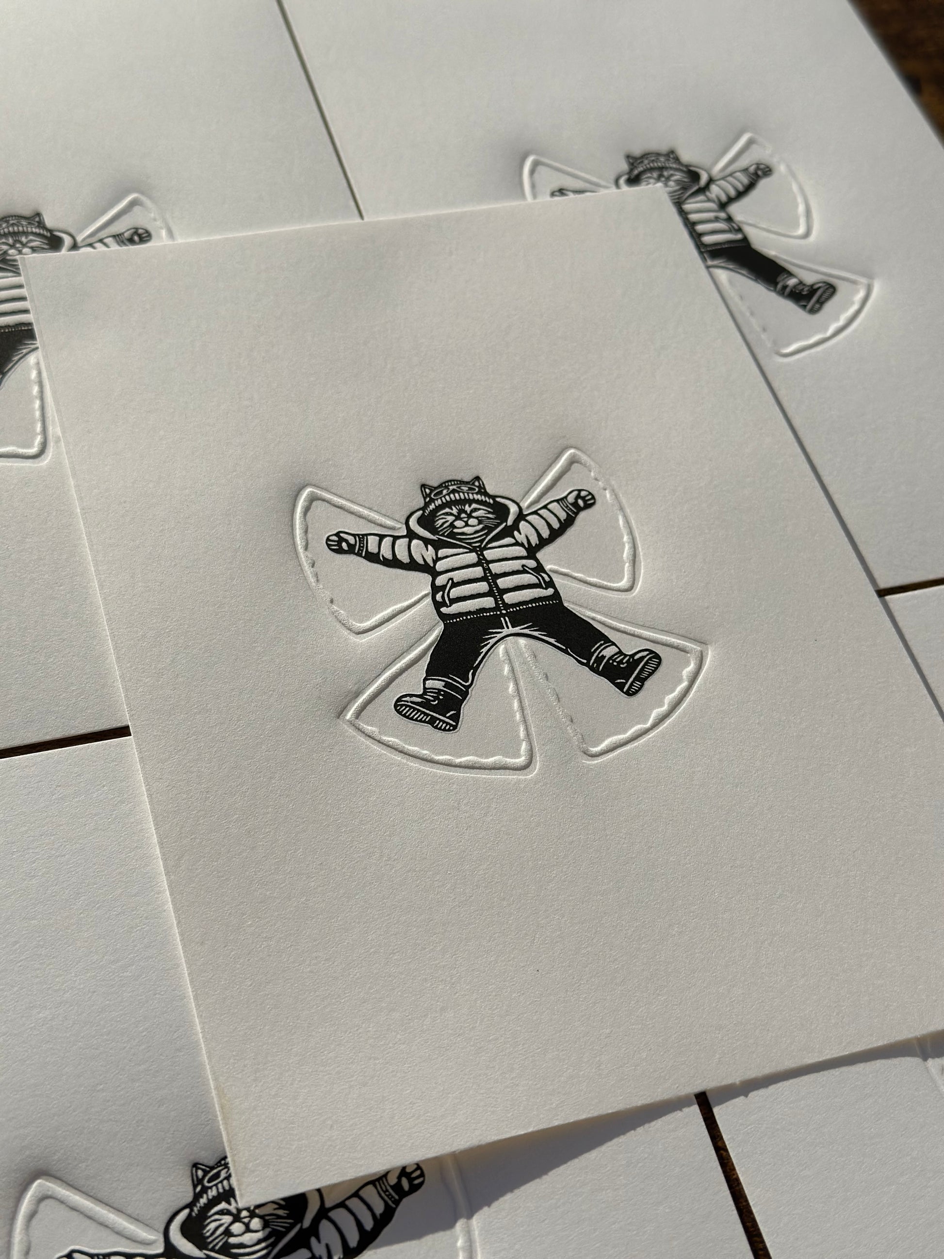 a black and white linocut print of a cat making a snow angel in the snow, the angel is embossed into the paper. 
