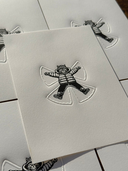 a black and white linocut print of a cat making a snow angel in the snow, the angel is embossed into the paper. 
