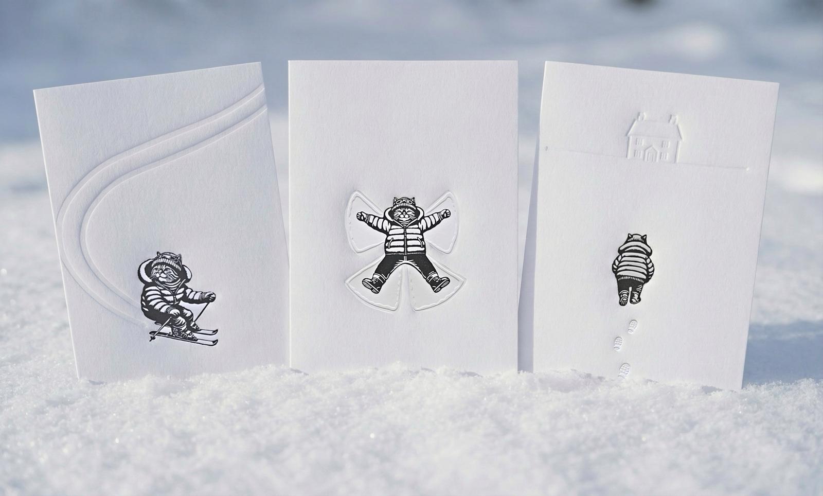 All three linocut prints of a cat, skiing, making snow angels and walking home through the snow, perfect gift for cat lovers, the three are situated in the snow. 