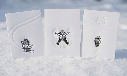 All three linocut prints of a cat, skiing, making snow angels and walking home through the snow, perfect gift for cat lovers, the three are situated in the snow. 