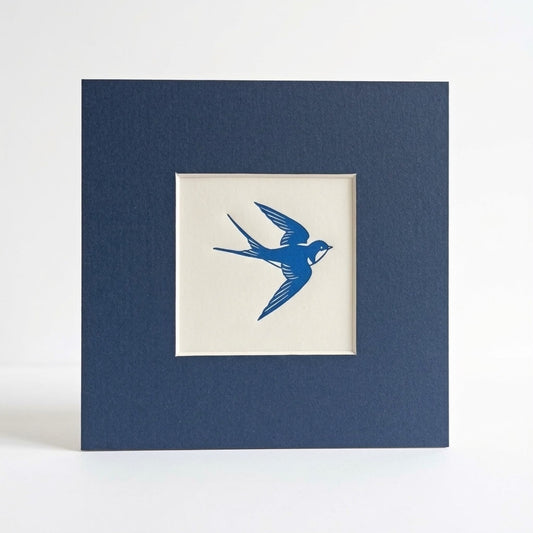 A linocut print of a swallow in blue in a navy blue mount photographed against a white background. 
