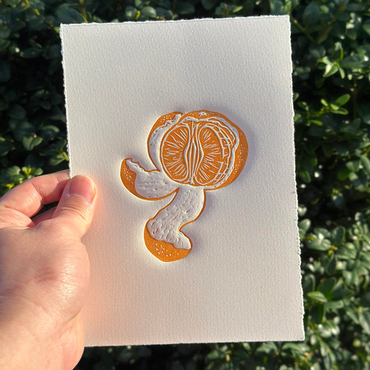 Linocut print of a partially peeled tangerine in vibrant orange on a white background, being held in someone's hand with foliage in the background