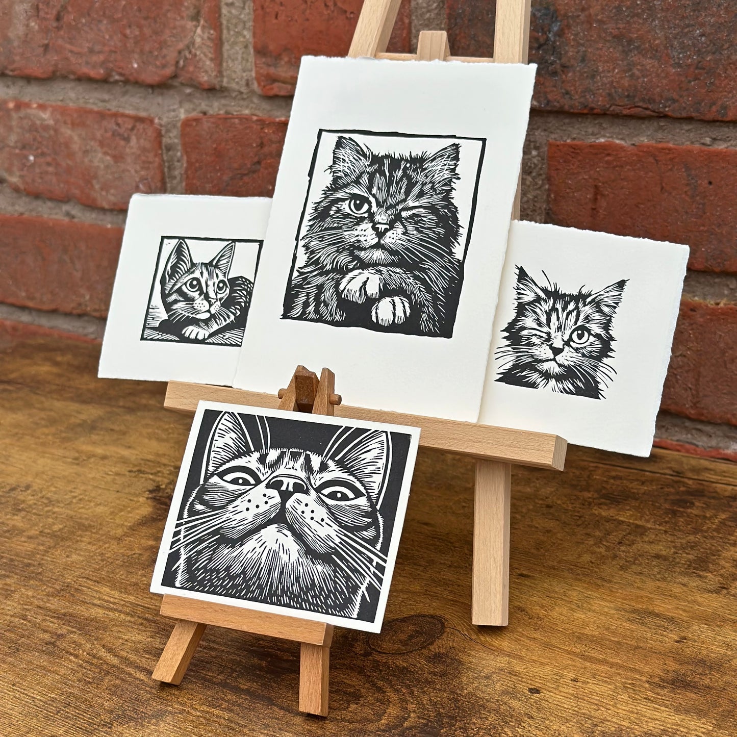 Set of four black and white cat linocut prints with a wooden easel against a brick wall.